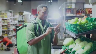 Market pazarında yiyecek arayarak akıllı telefon uygulaması kullanarak yeşil üniforma giyen genç bir siyah teslimatçı. Yiyecek dağıtımı.