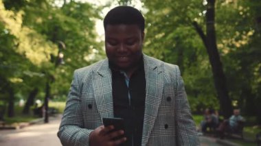 Tombul, gülümseyen, şık ceketli Afro-Amerikan bir adam sokakta akıllı telefonunu aktif olarak kullanıyor. Çevrimiçi olmak, sosyal ağlar, internette gezinmek. Erkek portresi, yakın görüntü, yavaş