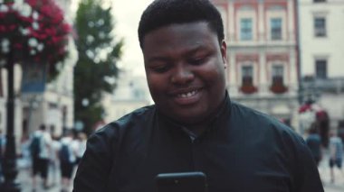 Şehir merkezinde akıllı telefonunu kullanarak yürüyen gülümseyen yakışıklı Afro-Amerikalı adamın yakın görüntüsü. İnternete, sosyal ağlara, aktif kullanıcılara bakıyorum. Instagram, yorum, blog. Erkek.