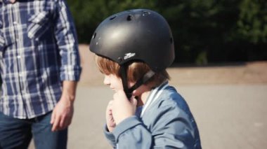 Kafasına siyah kask takmış güzel bir Slav çocuk, parkta bisiklet sürmeye başlıyor. Yakışıklı babacığına yardım et evlat. Yaz zamanı. Açık havada..