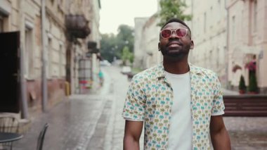 Yağmurdan sonra sokakta yürürken şık bir tişört giyen yakışıklı bir Afrikalı. Hoş görünüşlü hippi gülümsüyor. Açık havada. Şehir.