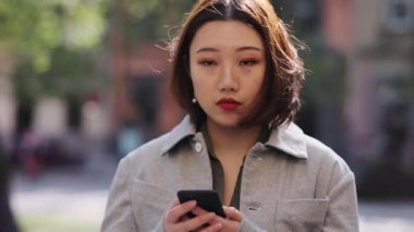 Ciddi bir portre genç Asyalı kadın telefon kullanıyor. Sokak kamerasına bak. İnternetten alışveriş yapıyor.