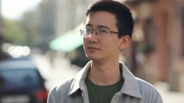 Asyalı ciddi bir adamın portresi, gözlüklü, sokak şehir merkezindeki kamera omuz silkerek güneş ışığına bakan sıradan, çekici, genç, neşeli, yakışıklı, yavaş çekimde başarılı, yakın çekim.