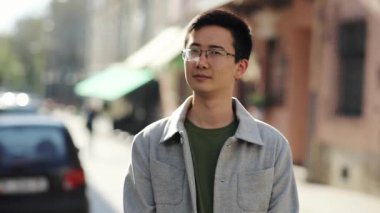 Gözlüklü yakışıklı Asyalı sokak şehir merkezindeki kamera omuz silkmelerine bakıyor. Gündelik modern, çekici, neşeli, genç ve yavaş çekimde başarılı bir portre. Yakın çekim.