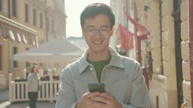 Gözlüklü genç Asyalı adam telefonda mutlu sokak şehir merkezinde günbatımında günbatımında modern, çekici, yakışıklı, başarılı, cep telefonlu, ağır çekimli Japon portresi.