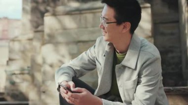 Gözlüklü Asyalı portre adam telefonda yan bakıyor. Şehir merkezinde güneş ışığı işinde gülümsüyor. Modern, çekici, neşeli, yakışıklı, yavaş çekimde başarılı.