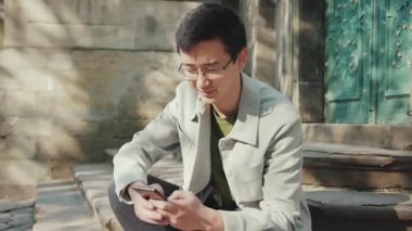 Gözlüklü mutlu Asyalı adam telefon kullanıyor ve kameraya bakıyor. Şehir merkezindeki güneş ışığına gülümsüyor. Modern, çekici, genç, neşeli, yakışıklı, yavaş çekimde başarılı.