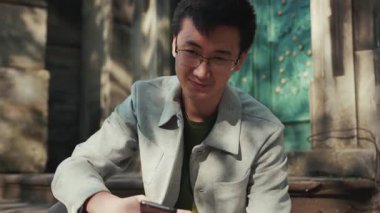 Kapalı, mutlu, gözlüklü Asyalı adam. Telefonunu kullan, kameraya bak. Şehir merkezindeki güneş ışığı işine gülümse.