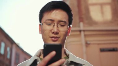 Gözlüklü Asyalı portre adamı telefonu kullan. Şehir merkezindeki kameraya bak. Güneş ışığı işi.