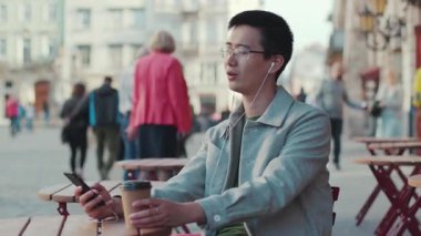 NEW YORK - 5 Nisan 2017: Genç Asyalı gözlüklü adam oturmuş müzik dinliyor şehir merkezinde gün batımında kahve içiyor modern çekici, yakışıklı ve başarılı cep telefonuyla yüzleş