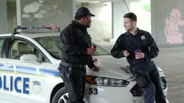 Gülümseyen iki polis memuru kahve içip iştahla çörek yerken devriye arabasının yanında konuşmak mutlu Afrika kökenli Amerikalı gangsterlerin üniforma giymesine yardımcı oluyor komik takım çalışması portresi yavaş çekim yakın çekim