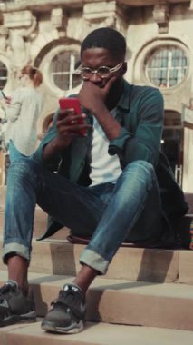 Etrafında canlı bir şehir ortamında merdivenlerde otururken akıllı telefonuna yoğun bir şekilde odaklanmış genç bir adam.