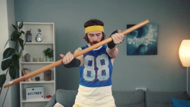 Renkli retro egzersiz kıyafetleri içinde bir fitness meraklısı evde egzersiz yaparken, sopayı dekor olarak kullanarak eğleniyor.