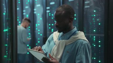 Çalışkan ve odaklanmış bir birey, çeşitli sunucularla çevrili modern bir veri merkezinde bir tablet kullanıyor.