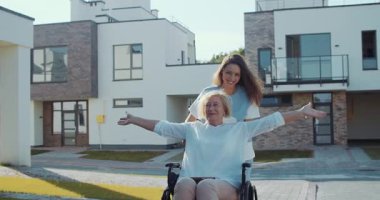 Tekerlekli sandalyedeki bir kadın, güzel ve modern evlerin dışında şefkatli bir bakıcı ile neşe içinde poz verir.