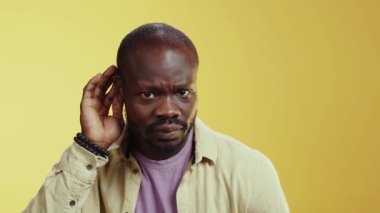 Parlak sarı bir ortamda, sakallı bir adam dikkatle dinleyen bir nesneyi inceliyor. Sıcak gülüşü anlayış gösteriyor. Tişörtlü bir figür onun yanında dururken.