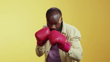 Kırmızı eldivenlerle boks teknikleri sergileyen bir adam, parlak sarı bir arka plana karşı, odaklanma ve yetenek sergiliyor. Beyaz gömlekli başka bir adam çeşitlilik katıyor.