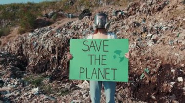 Bir aktivist, çevresel atık ve kirliliğin ortasında gezegeni kurtarmak için harekete geçmeye çağıran parlak bir işaret tutuyor.