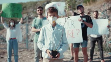 Genç bir aktivist, canlı bir iklim protestosunda küçük bir bitki tutuyor ve arka planda destekçiler görülüyor.
