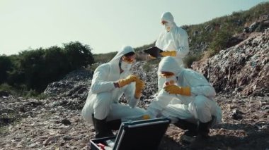 Koruyucu giysiler giymiş bir grup araştırmacı bir açık hava alanında çeşitli örnekler inceliyor.