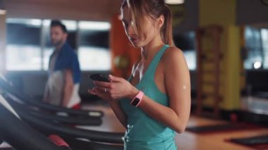 Koşu bandındaki genç bir kadının portresi spor salonunda güneş ışığı spor spor fitness internet yüz teknolojisi yaşam tarzı spor spor spor kıyafetleri vücut egzersizi ağır çekim akıllı telefon.