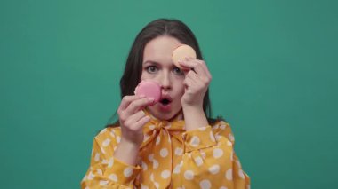 Komik suratlı komik genç bir kadın esmer yeşil bir arka planda iki renkli badem kurabiyesiyle komik bir şekilde el kol hareketi yapıyor. Mutlu, neşeli bir ruh hali. Lezzetli, tatlı düşkünü. Yavaş çekim, yakın görüntü, kadın portresi.