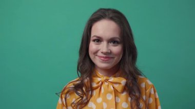 Kıvırcık saçlı neşeli genç bir kadın yeşil arka planda duruyor, doğrudan kameraya bakıyor ve neşeli bir şekilde gülümsüyor. Pozitif duygular, gerçek duygular, iyi hissetmek. Ağır çekim, yakın çekim, kadın portresi.