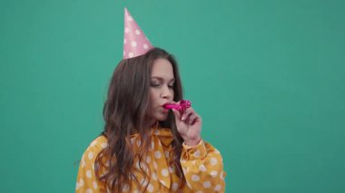 Doğum günü şapkalı çekici kıvırcık kadın yeşil arka planda, doğum günü kornası kullanıyor. Eğlenmek, doğum günü partisi, neşeli bir ruh hali. Yavaş çekim, yakın görüntü, kadın portresi.