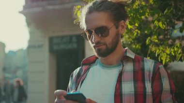 Güneş gözlüklü genç adam telefon sokağından günlük kıyafet şehirlerine mesaj atıyor güneşli, şık, zarif, mükemmel görünümlü yavaş çekimde portreye yakından bakıyor.