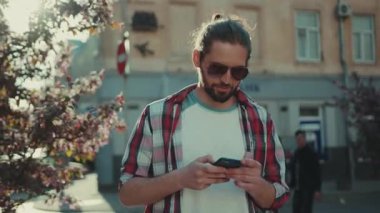 Güneş gözlüklü güler yüzlü genç adam telefon caddesinde yürüyor günlük kıyafet şehirleri geziyor güneşli güneş ışığına bakıyor genç mükemmel görünümlü yavaş çekim portresi yakın çekim