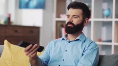 Bakımlı sakallı bir adam rahat bir kanepede oturup uzaktan kumandayla televizyon seyrediyor.