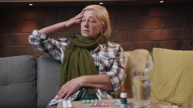 Başı ağrıyan yaşlı bir kadının portresi. Kanepede oturmuş sıcaklığı ölçüyor. Hasta, mutsuz, yaşlı, yaşlı, yaşlı, yaşlı, yaşlı, yaşlı, yaşlı, yaşlı, emekli, üzgün bir kadın.