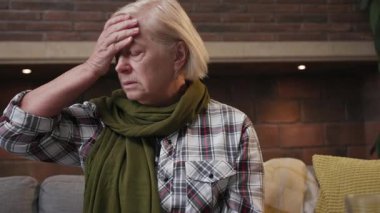 Baş ağrısı olan yaşlı bir kadının hasta portresi kanepede oturan sıcaklığı ölçer. Yaşlı, mutsuz, yaşlı, yaşlı, yaşlı, yaşlı, yaşlı, yaşlı, yaşlı, emekli, emekli, ağır çekimde emekli olur.