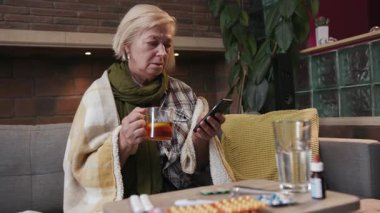 Yaşlı kadın çay içer telefonda konuşur evde ateşlenir hasta, hasta, soğuk algınlığı hastası, virüs portresi yavaş çekimde hapları kapatır.