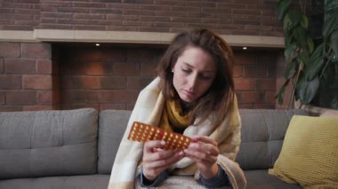 Baş ağrısı çeken genç bir kadın kanepede oturup çay içiyor. Virüs hastalığı, soğuk hava sıcaklığı semptomları. Kanepe enfeksiyonu ilaçları.