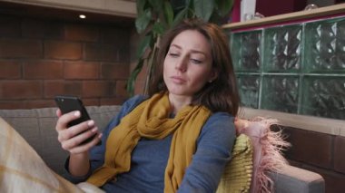 Mutsuz bir kadın kanepede uzanıyor ve cep telefonunu kullanıyor. Çay virüsü hastalığı, soğuk hastalık belirtileri, sağlık portresi battaniye enfeksiyonu haplarını yavaş çekimde kapıyor.