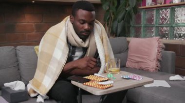 Üzgün Afro-Amerikalı genç adam kanepede oturuyor. Evde ateş hapları var. Çay içiyor. Mutsuz, soğuk algınlığı hastası, hastalıklı, hasta, virüs alerjisi olan biri.