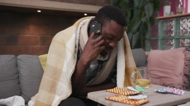 Afro-Amerikalı genç adam koltukta oturmuş telefonda konuşuyor. Evde çay içiyor. Üzgün, grip, hasta, soğuk algınlığı hastası, virüs alerjisi olan biri.