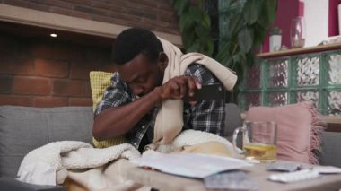 Afro-Amerikalı genç adam kanepede uzanmış, öksürüyor, evde çay içiyor, üzgün, grip, hasta, soğuk algınlığı hastası, virüs alerjisi, yavaş çekimde portreyi kapatıyor.