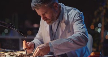 Profesyonel yetişkin Kafkasyalı tamirci karanlık ofis laboratuarında bozuk İHA cihazı için teknik tamir devresi lehimliyor. Mühendislik,