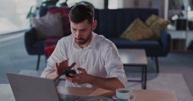 Bir adam şık bir ofis ortamında dizüstü bilgisayar üzerinde çalışırken dikkatle akıllı telefonuna odaklanır.