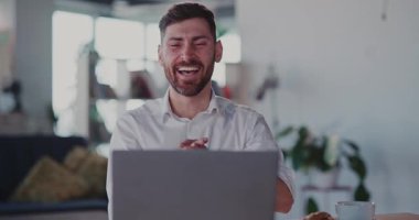 Komik sakallı yetişkin beyaz adam dizüstü bilgisayar kullanıyor eğlenceli videolar seyrediyor ve şık bir dairede oturup gülüyor..