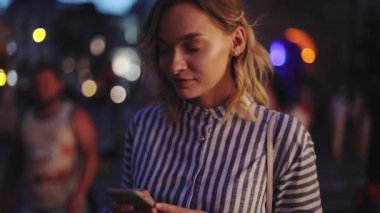 Yavaş çekim yakın çekim çekici genç kadın gece yürüyüşü sırasında telefon tebessümü kullanarak kadın kız kıza iletişim mutlu kasaba ışığı teknolojisi akşam portresi tutun.