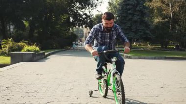 Parkta çocuklar için yeşil bisiklet süren kot pantolon ve ekose gömlek giyen oyuncu bir adam. Neşeli baba çocukluğu hatırlar. Yaz zamanı. Açık havada..