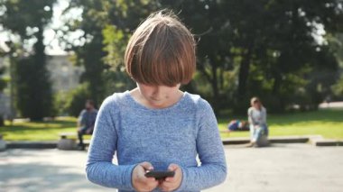 Mavi kazak giyen küçük beyaz çocuk elinde akıllı telefonu tutuyor, kavga ediyor. Elinde aletle caddede yürüyen tatlı bir çocuk. Şehre. Açık havada..