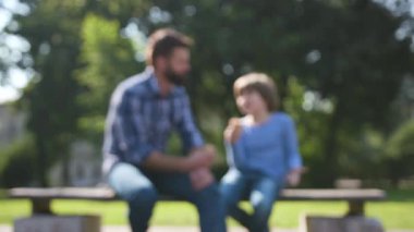 Koyu saçlı, yakışıklı beyaz bir adam yakışıklı oğlunun yanında oturuyor ve konuşuyor. Parkta oynadıktan sonra dondurma yemek güzeldi. Açık havada. Yaz zamanı.