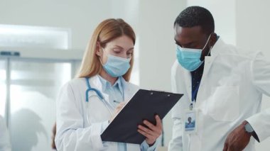 Çok ırklı sağlık doktorları koridorda iletişim kuruyor. Kendini izole etmek için ameliyat maskesini takıyorlar. İşbirliği. Pandemik koronavirüs konsepti.