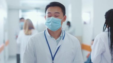 Beyaz tıbbi önlük ve cerrahi maske takmış genç Çinli doktorun portresi koridor hastanesindeki kameraya bakıyor. Tıbbi personel. Coronavirus karantinası. Kamu güvenliği. Sağlık Hizmetleri.