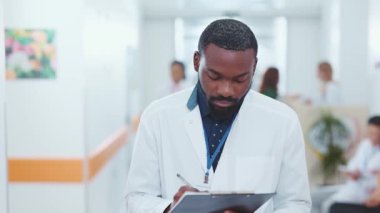 Afrikalı Amerikalı yakışıklı doktor terapistin portresi. Klinik kayıtlar yazıyor. Koridordaki hastanede kameraya bakıyor. Tıbbi personel. Çalışma alanı, sağlık ortamı.