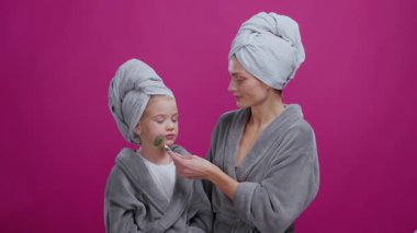 Anne ve kızı banyoda cilt bakımı yapıyorlar. Mutlu, rahatlatıcı, komik görünümlü bir çocuğun anne masajı. Mor arka plan. Güzellik konsepti.